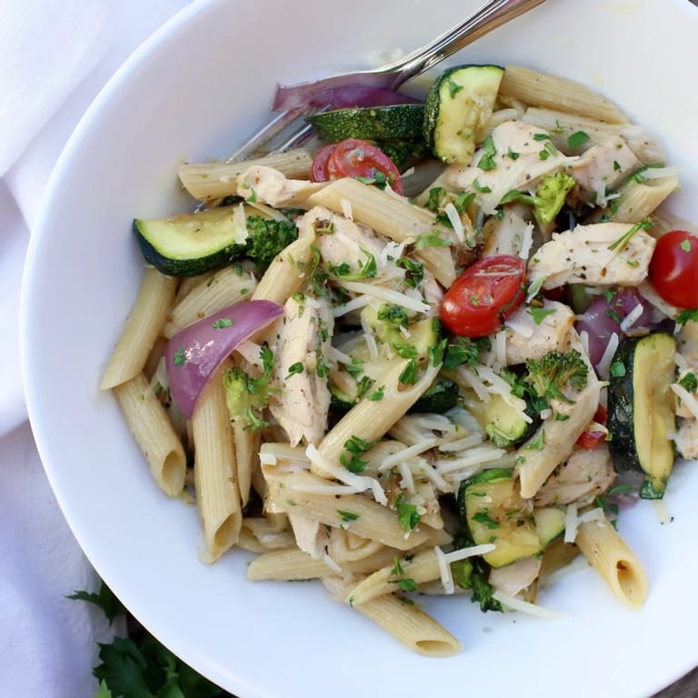 A close-up of garlic herb chicken, red peppers, zucchini, and cherry tomatoes mixed with penne on a white plate, ready to serve.
